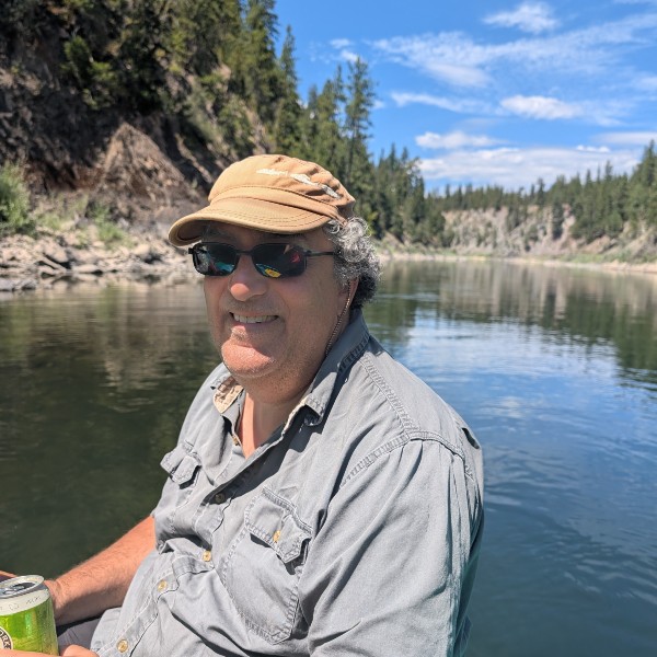 Bob Jaffe floating on water in the mountains wearing a baseball cap and sunglasses smiling at the camera.