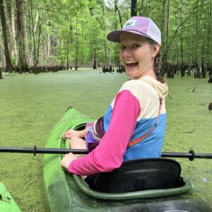 Lily Amato Smiling at the camera while kayaking through water covered in Duckweed and surrounded by trees.