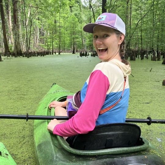 Lily Amato Smiling at the camera while kayaking through water covered in Duckweed and surrounded by trees.
