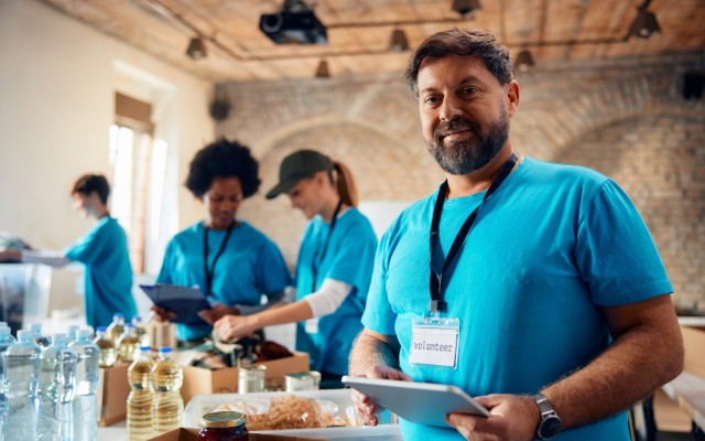 A man in a blue shirt holding a tablet with a tag around his neck saying "volunteer" smiling at the camera with others in similar outfits in the background.