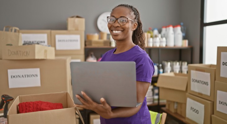 A person holding a laptop and smiling at the cameral while surrounded by boxes.