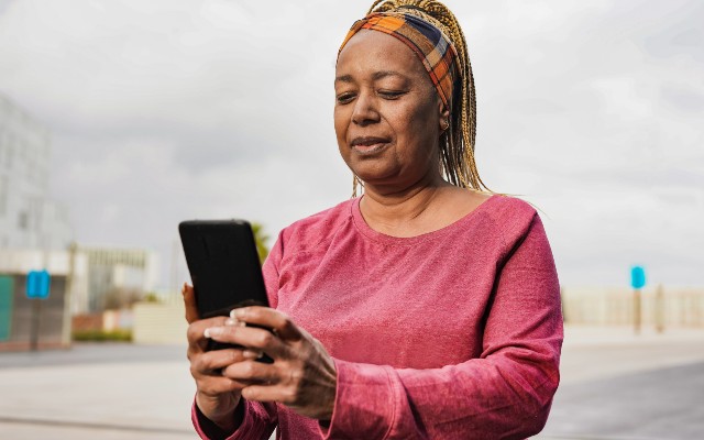 An older woman in a pink shirt standing outside and looking at her phone.