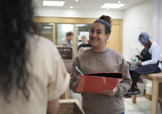A woman smiling at another woman to the left of the screen while holding a binder and pen.