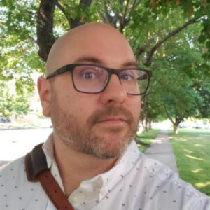 Bill looking at the camera while walking on a sidewalk near grass and trees.